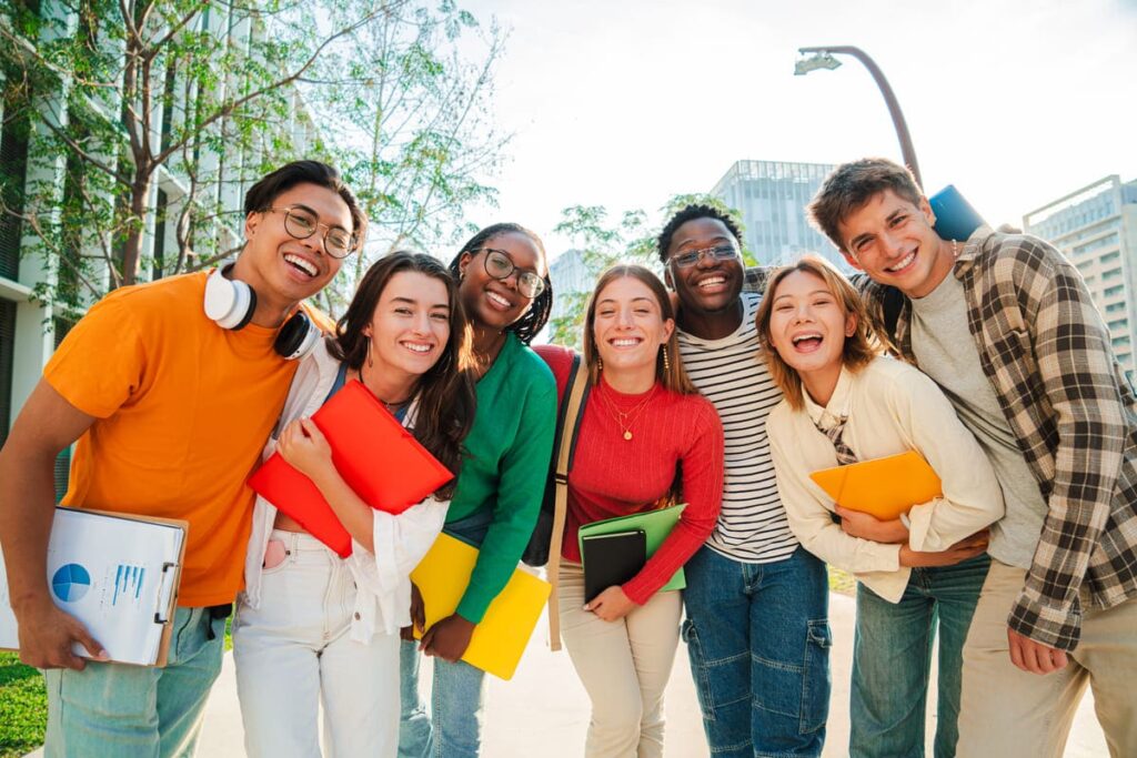 Groupe de sept jeunes adultes souriants debout à l'extérieur tenant des cahiers et des dossiers, avec des arbres et des bâtiments en arrière-plan.