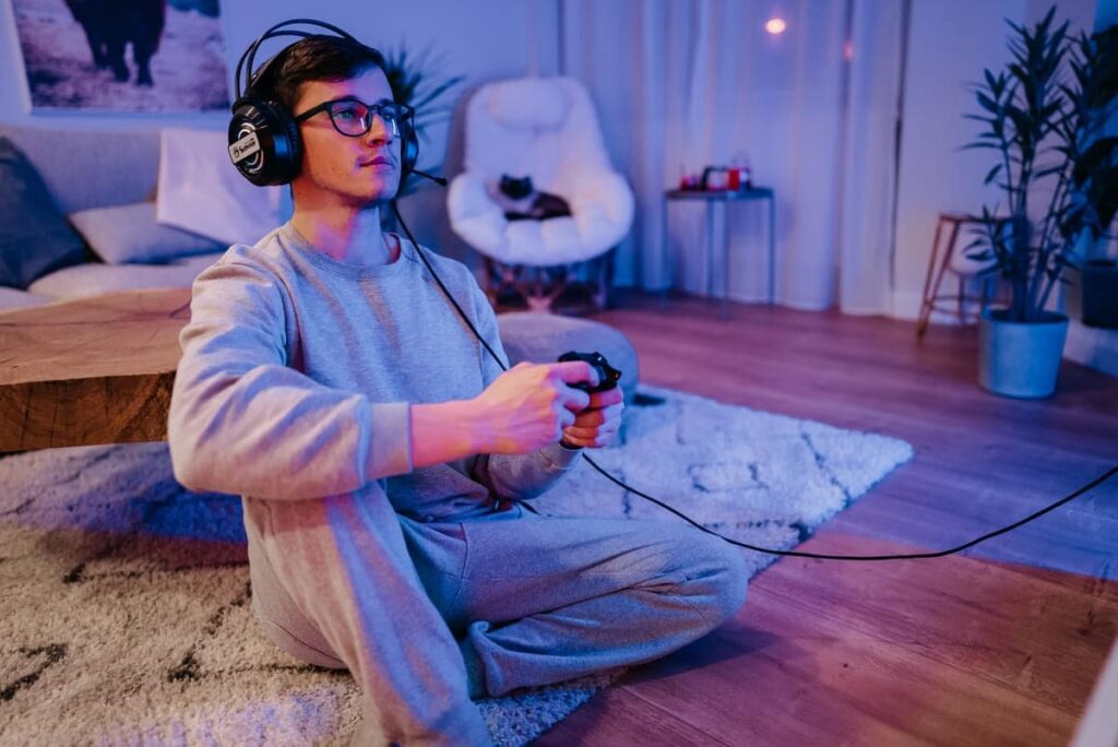 Une personne assise sur un tapis dans un salon, portant des écouteurs et jouant à un jeu vidéo avec une manette. Un chat se repose sur une chaise en arrière-plan.