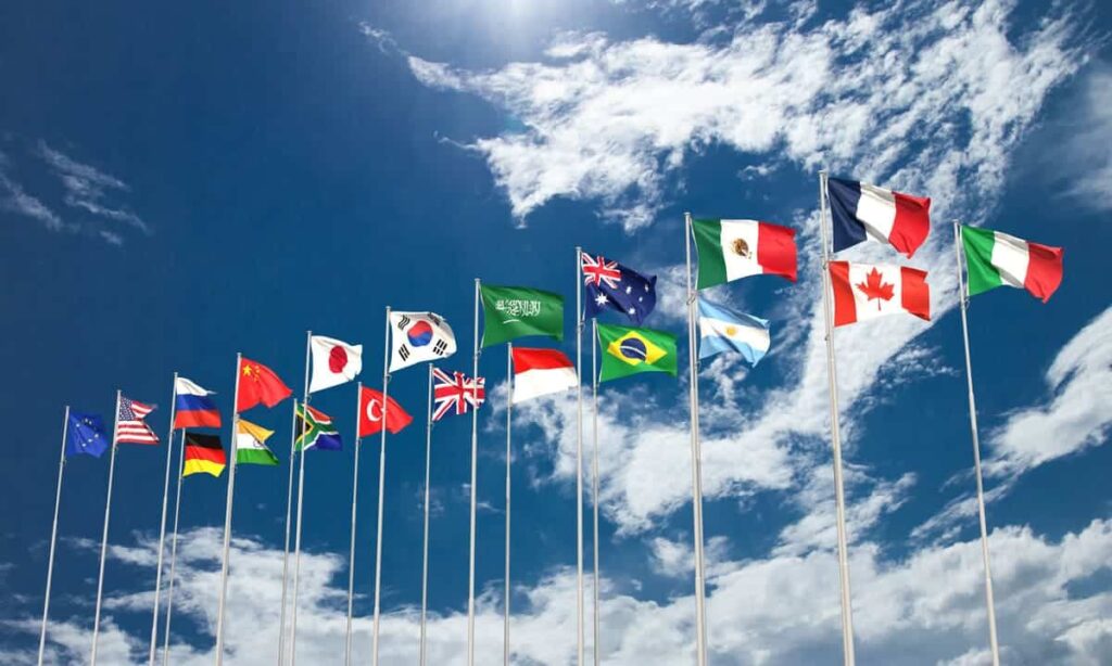 Les drapeaux nationaux de divers pays, dont les États-Unis, la Chine, l'Allemagne, le Japon et d'autres, sont affichés sur des mâts contre un ciel bleu parsemé de nuages.