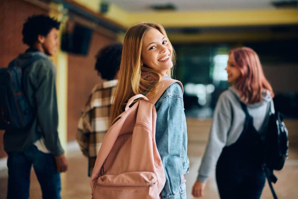 Une jeune femme souriante aux longs cheveux blonds et au sac à dos rose regarde en arrière tout en marchant avec trois amis dans un couloir.