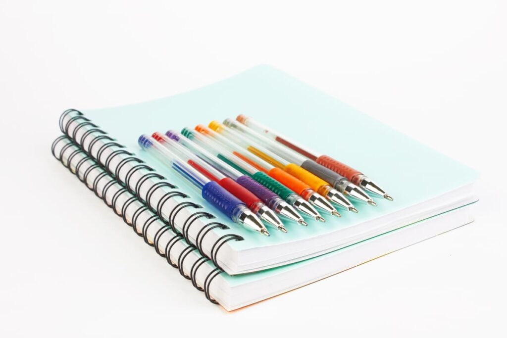 Deux cahiers à spirales avec des couvertures colorées sont empilés ; dix stylos à bille avec des poignées de couleurs différentes sont posés en diagonale sur le cahier supérieur.