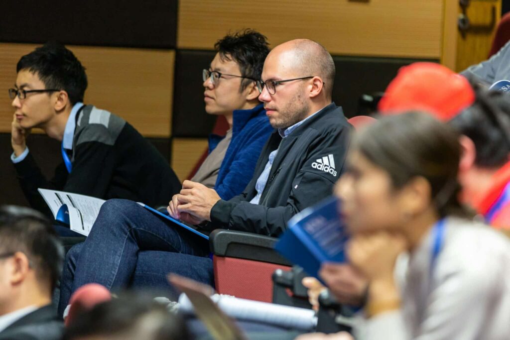 Un groupe de personnes assises dans un auditorium, écoutant attentivement une présentation. Certains tiennent des papiers ou des cahiers.