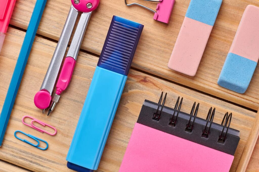 Assortiment de fournitures de bureau comprenant un surligneur bleu, un compas, des gommes, un trombone, des trombones et un bloc-notes en spirale avec du papier rose sur une surface en bois.