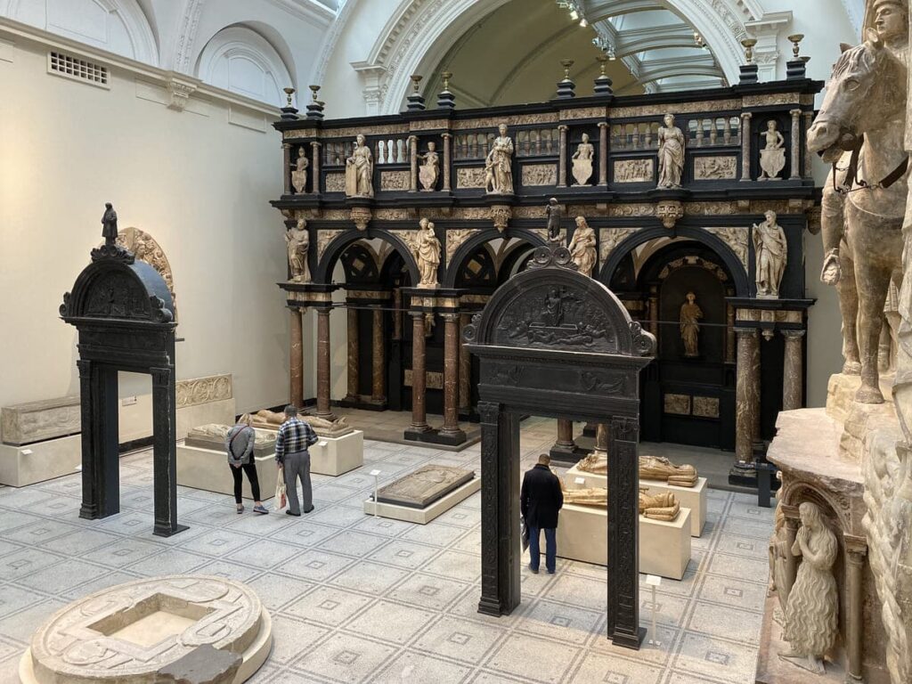 Les gens observent les expositions dans une galerie de musée, présentant des sculptures classiques, des objets en pierre et une structure architecturale ornée à l'arrière.