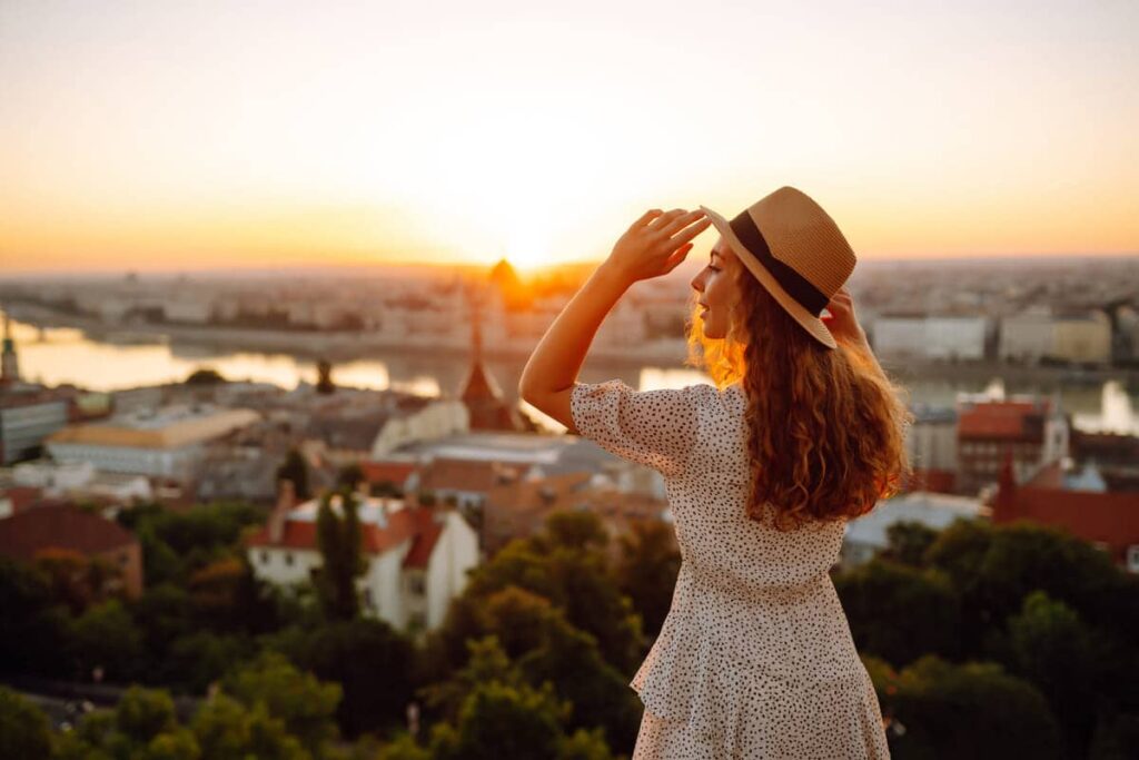 Une femme portant un chapeau et une robe à pois se tient sur une colline surplombant un paysage urbain au coucher du soleil, se protégeant les yeux d'une main.