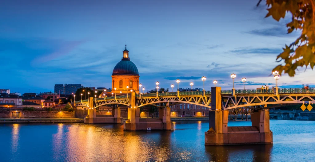 Un pont illuminé enjambe une rivière calme au crépuscule, avec un bâtiment en forme de dôme en arrière-plan. Le ciel est partiellement nuageux.