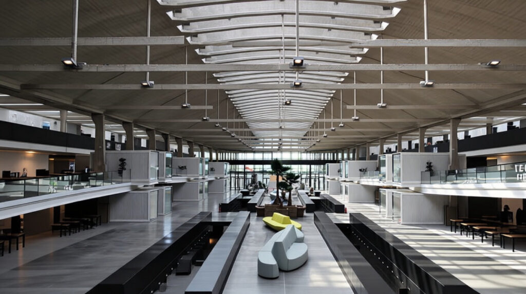 Bureau moderne et spacieux avec de hauts plafonds, de grandes fenêtres, plusieurs bureaux et un coin salon central avec des meubles contemporains.