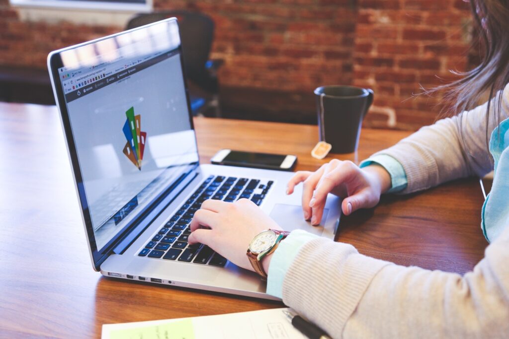 Personne travaillant sur un ordinateur portable avec un graphique coloré à l'écran, un smartphone, du thé et des papiers sur une table en bois dans un bureau aux murs de briques.